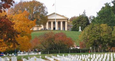Arlington House, a Greek Revival style mansion on a hill, with a section of grave markers at Arlington National Cemetery in the foreground. Trees on the property are showing fall colors of green, red, and orange. The American flag in front of house is at half-staff.