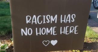 Lawn sign, brown with white lettering, says "Racism has no home here" with two hearts