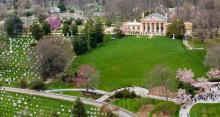 An aerial view of Arlington House, lawn, and part of the cemetery in the spring time. Cherry blossom trees are in bloom and the flag is at half mass.