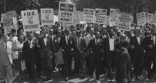 Civil rights leaders march down the middle of the street with crowds of people behind them holding signs advocating for voting rights, jobs for all, full employment, integrated schools, and equal rights.