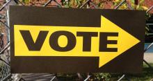 Brown sign on chain link fence with large yellow arrow that says "Vote."